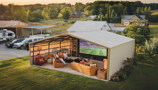 The image depicts a spacious, enclosed metal carport transformed into a vibrant sports viewing lounge, featuring a family or group of friends enjoying a game day in comfortable seating with a large screen displaying a sports event, set in a picturesque rural or suburban environment with various roof styles, parked vehicles, an RV, and lush greenery, all highlighted by a warm and inviting color palette to appeal to homeowners, rural landowners, small business owners, and RV enthusiasts across the southern, central, and western United States.
